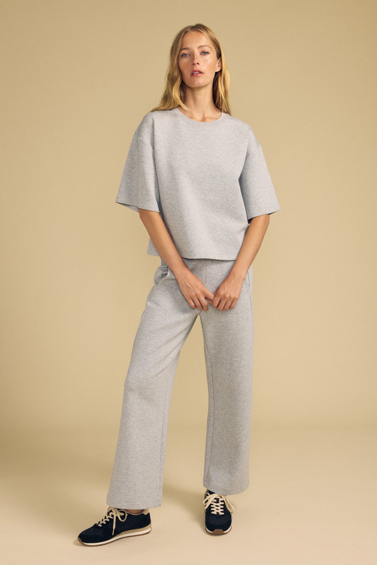 A woman stands against a beige backdrop wearing Velvet by Graham & Spencer's ARA SWEATPANT, a pair of loose-fitting grey wide-leg pants, styled with a matching short-sleeve top and black sneakers with white laces.