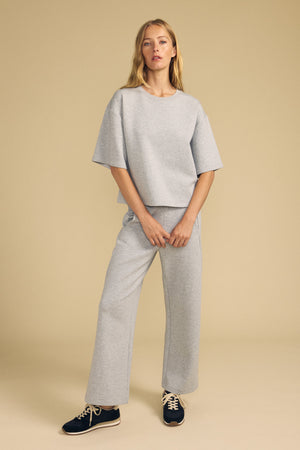 A woman stands against a beige backdrop wearing Velvet by Graham & Spencer's ARA SWEATPANT, a pair of loose-fitting grey wide-leg pants, styled with a matching short-sleeve top and black sneakers with white laces.