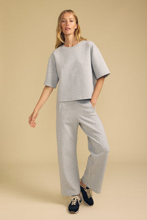 A woman stands against a beige background wearing the Velvet by Graham & Spencer ARA SWEATPANT—a pair of loose-fitting, light gray wide-leg pants—paired with a matching short-sleeve top and black sneakers.