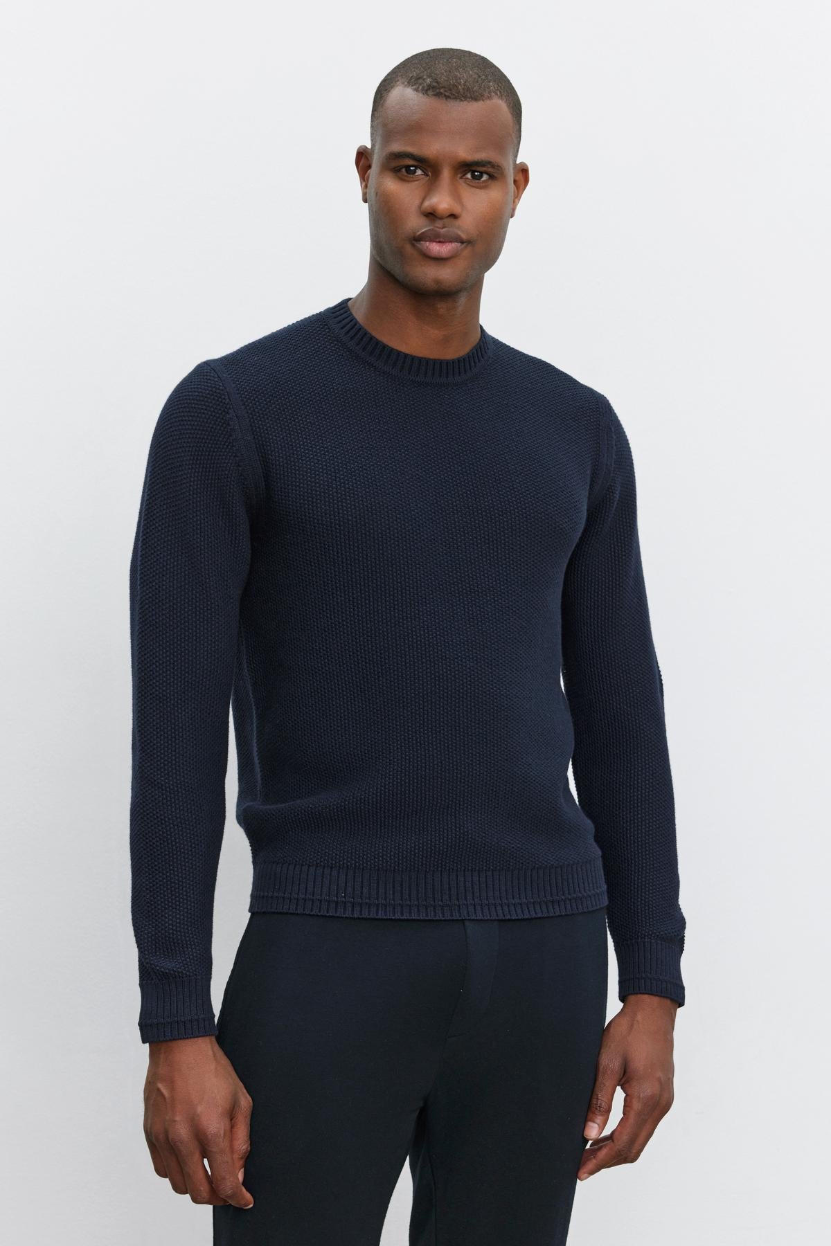   A man stands against a plain background, wearing the MARCO SWEATER from Velvet by Graham & Spencer, made of a navy blue cotton cashmere blend with a seed stitch texture, paired with black pants. He has a neutral facial expression and his hands are relaxed by his sides. 