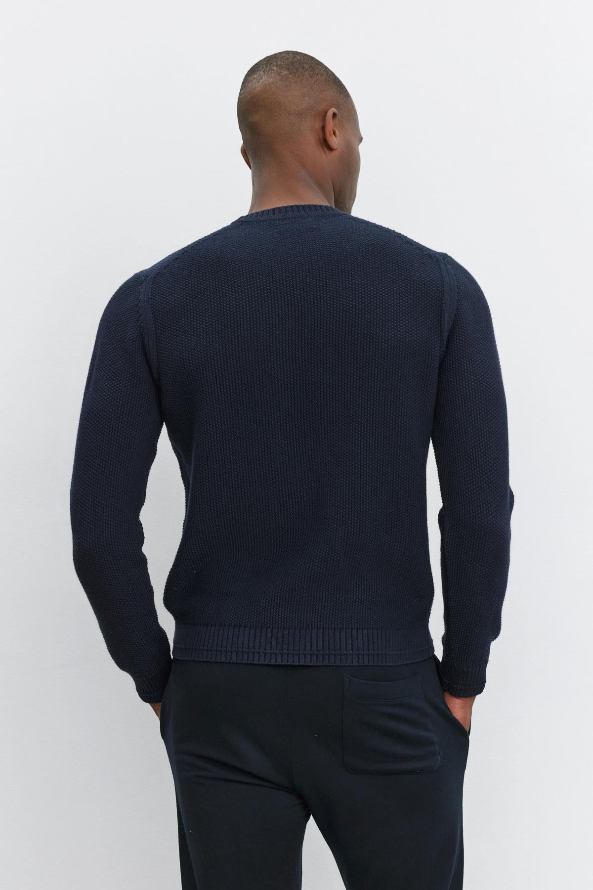   A man is shown from the back, wearing Velvet by Graham & Spencer's MARCO SWEATER—a long-sleeved, dark-colored sweater with a seed stitch texture—and dark pants against a plain background. 