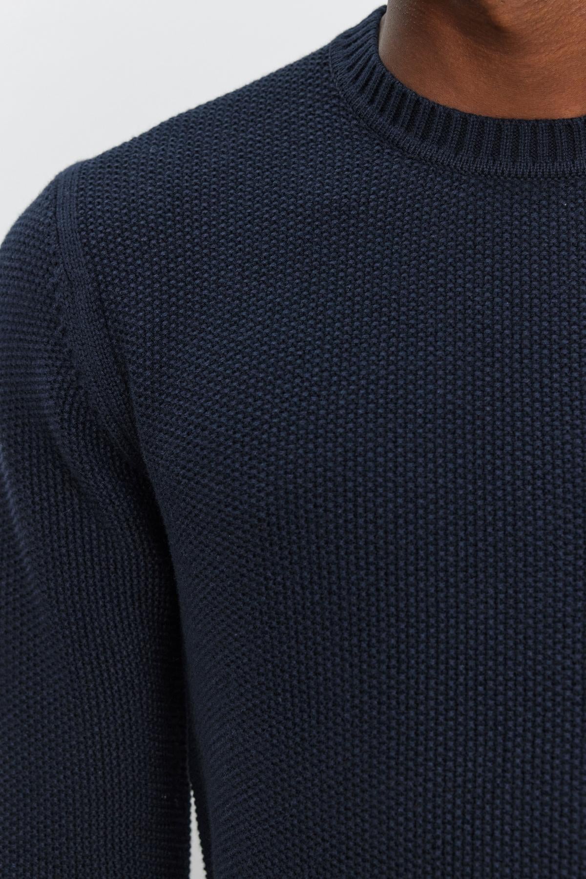   Close-up of the upper torso of a person wearing the MARCO SWEATER, a textured navy blue knit by Velvet by Graham & Spencer featuring seed stitch detailing. Only the sweater and a partial view of the person's neck and jawline are visible. 