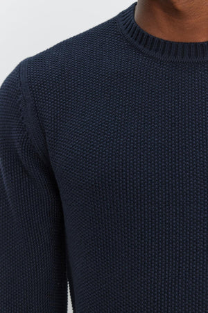 Close-up of the upper torso of a person wearing the MARCO SWEATER, a textured navy blue knit by Velvet by Graham & Spencer featuring seed stitch detailing. Only the sweater and a partial view of the person's neck and jawline are visible.