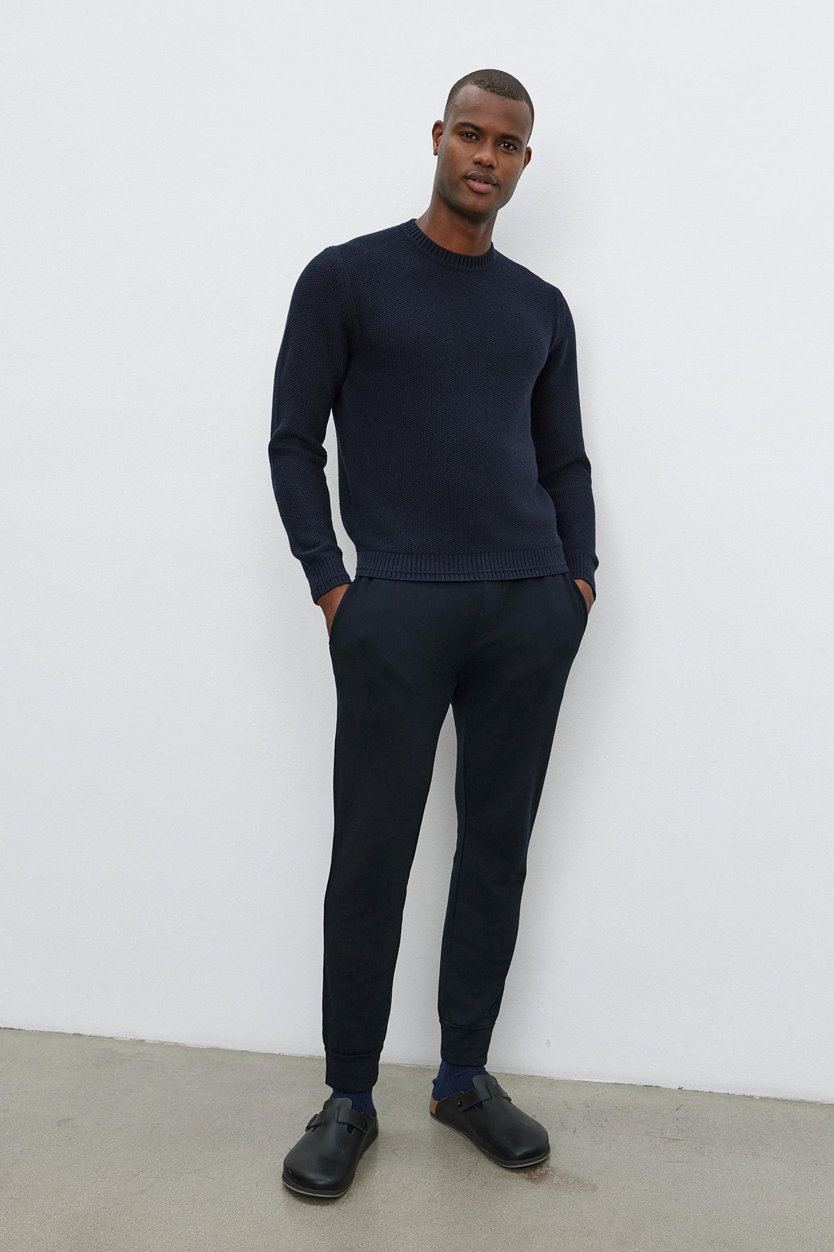   A person stands against a white wall, wearing the navy MARCO SWEATER with a seed stitch texture by Velvet by Graham & Spencer, black pants, and black shoes, with hands in pockets. 