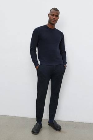 A person stands against a white wall, wearing the navy MARCO SWEATER with a seed stitch texture by Velvet by Graham & Spencer, black pants, and black shoes, with hands in pockets.