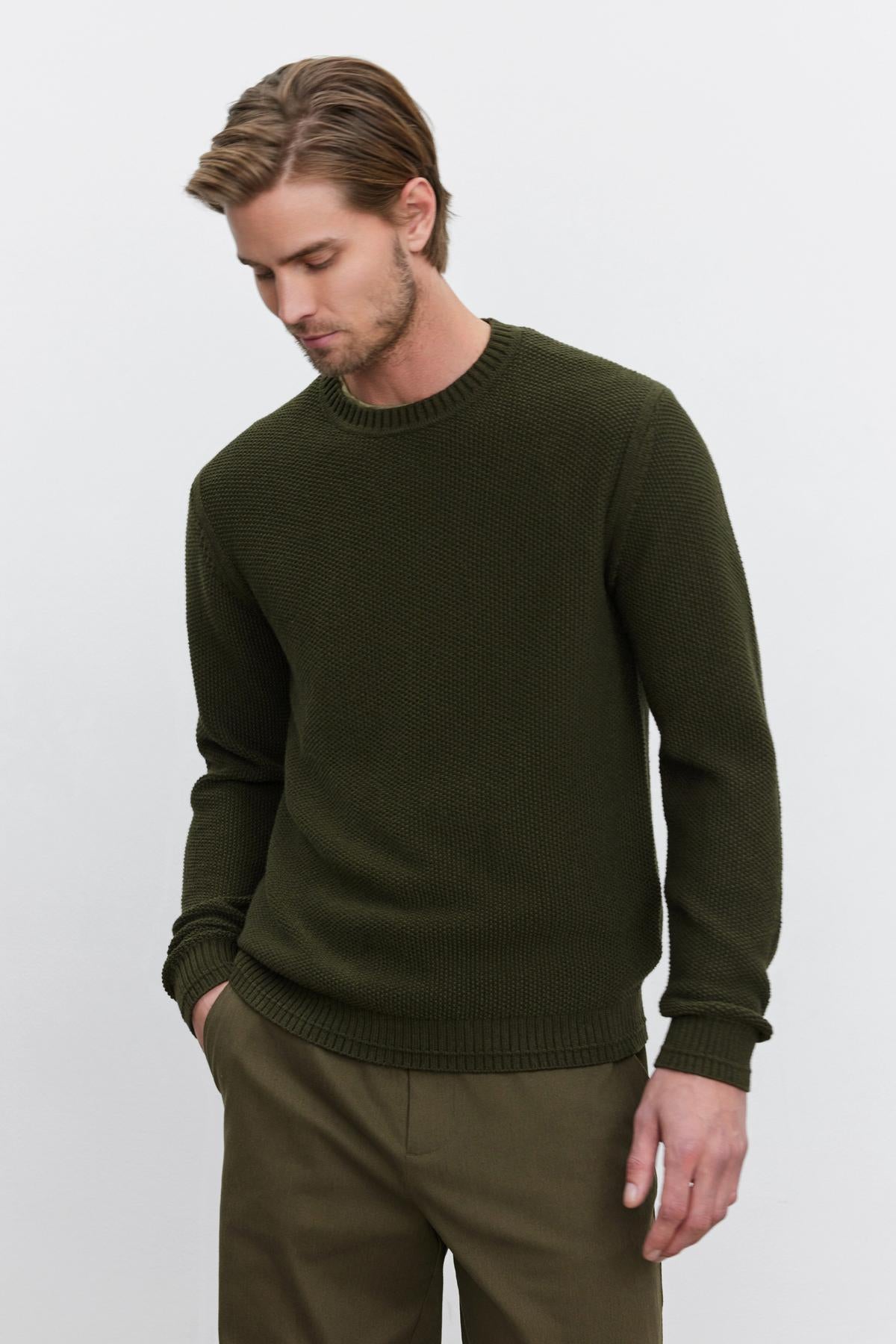   A man with light brown hair, wearing the MARCO SWEATER by Velvet by Graham & Spencer, made of a cozy cotton cashmere blend in green, and olive green pants, stands with one hand in his pocket, looking slightly down and to the side. 