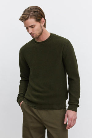 A man with light brown hair, wearing the MARCO SWEATER by Velvet by Graham & Spencer, made of a cozy cotton cashmere blend in green, and olive green pants, stands with one hand in his pocket, looking slightly down and to the side.