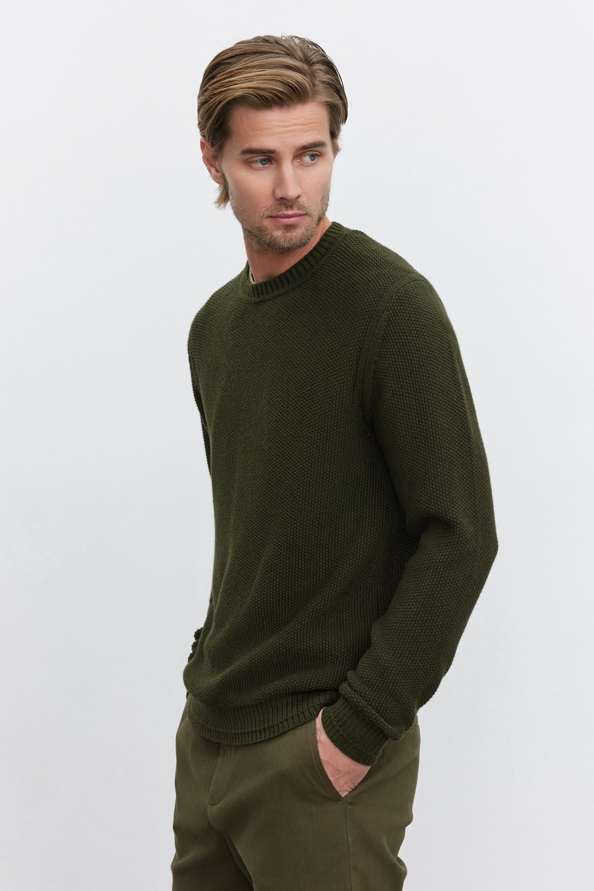   A man wearing the MARCO SWEATER by Velvet by Graham & Spencer, crafted from a dark green cotton-cashmere blend with seed stitch texture, and olive pants, stands against a neutral background. He has his hands in his pockets and is looking off to the side. 