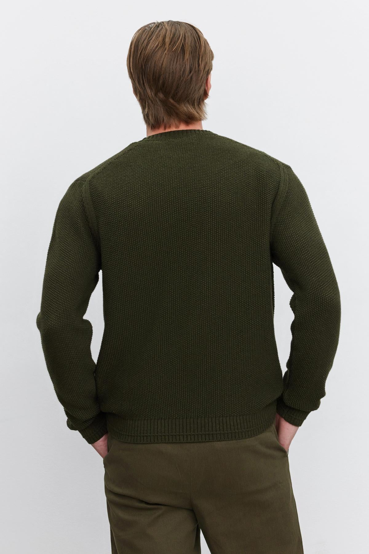   A person is standing with their back to the camera, wearing the MARCO SWEATER from Velvet by Graham & Spencer, a dark green knitted sweater featuring a seed stitch texture, paired with matching green pants. 