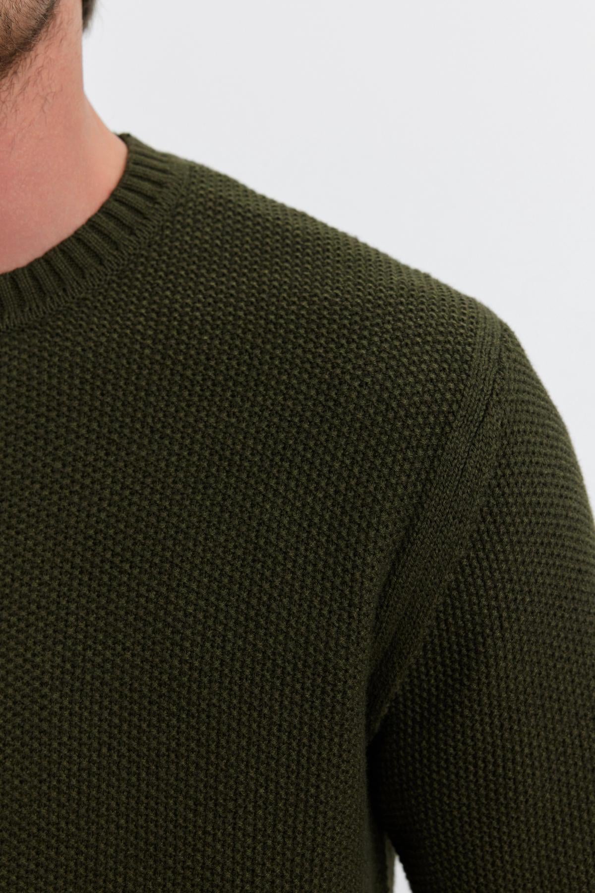   Close-up of a person wearing the MARCO SWEATER, a textured, dark green piece with a seed stitch pattern. The image highlights the shoulder, sleeve, and part of the neckline crafted from a soft cotton cashmere blend by Velvet by Graham & Spencer. No additional details of the person are visible. 