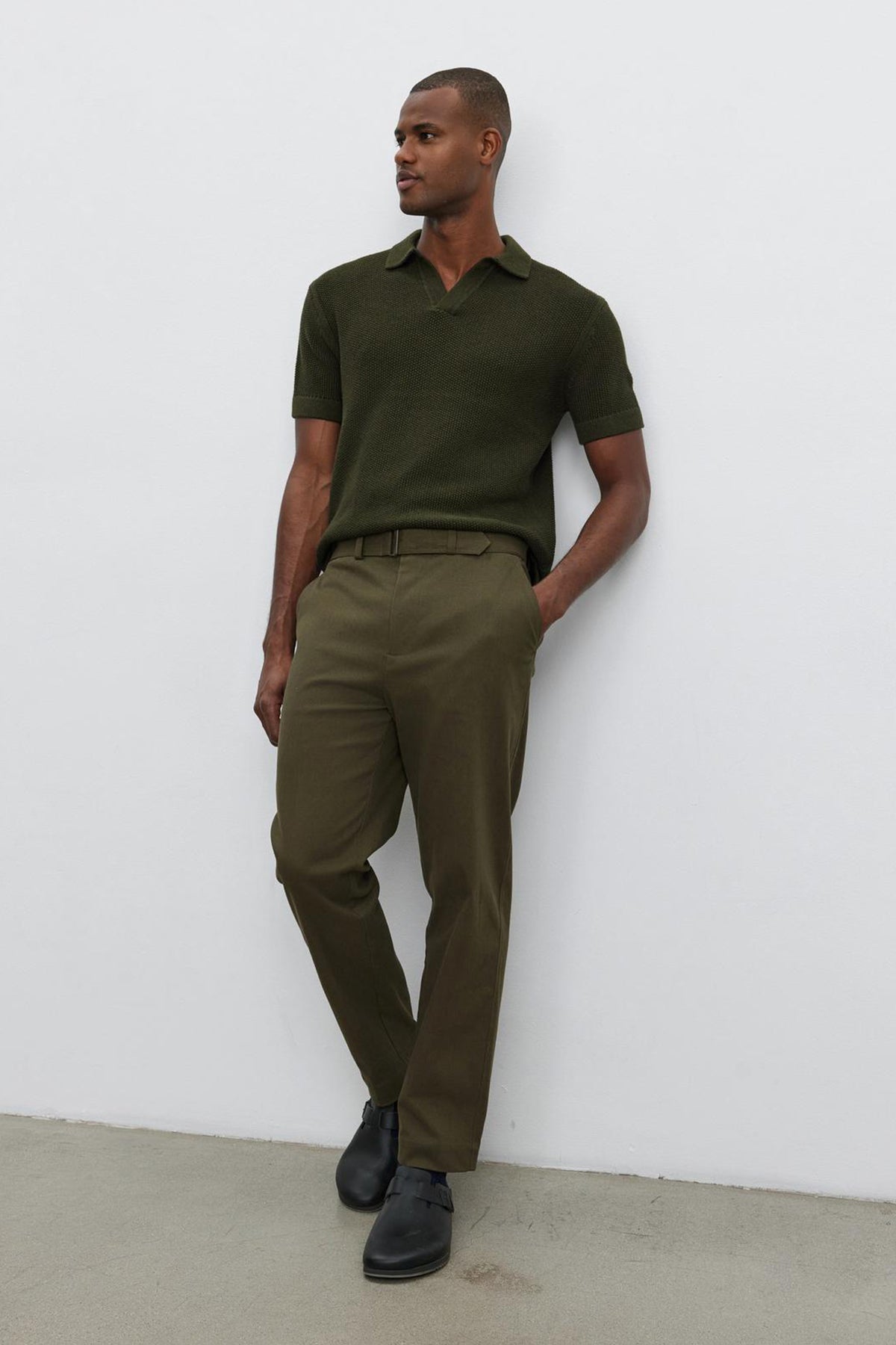   Person standing against a white wall, wearing the Velvet by Graham & Spencer NIKLAS SWEATER POLO in olive green with a subtle seed stitch texture, matching olive green pants, and black shoes. 