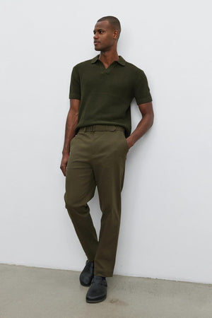 Person standing against a white wall, wearing the Velvet by Graham & Spencer NIKLAS SWEATER POLO in olive green with a subtle seed stitch texture, matching olive green pants, and black shoes.