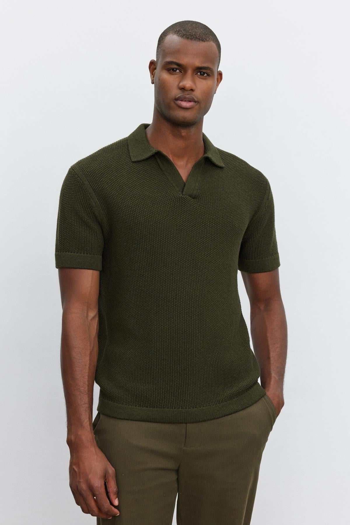   A man wearing the NIKLAS SWEATER POLO by Velvet by Graham & Spencer in an olive green hue, featuring a seed stitch texture, paired with matching olive green pants, standing with his hands partially in his pockets. 