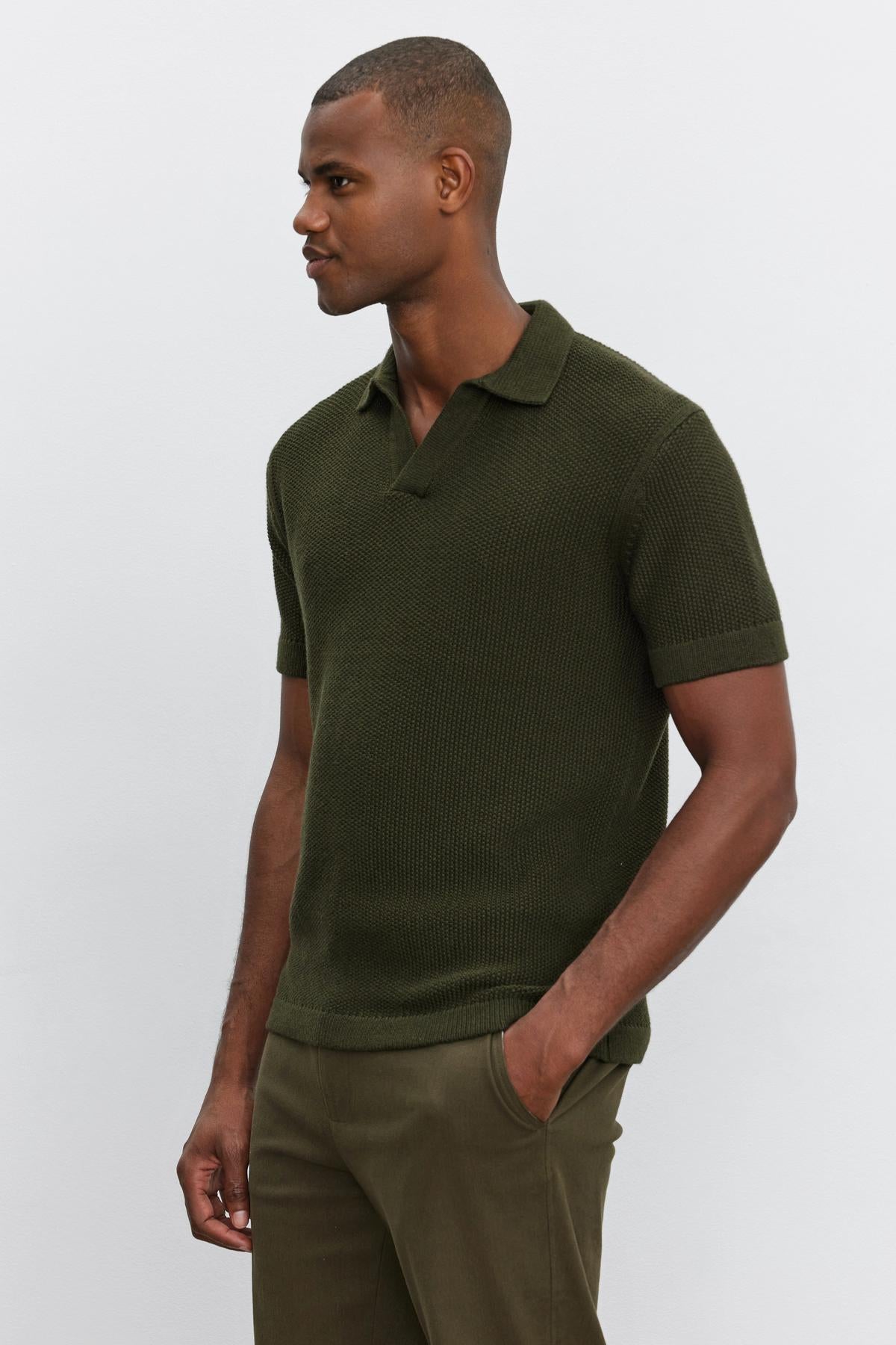   A person is standing against a plain background, wearing the dark green NIKLAS SWEATER POLO from Velvet by Graham & Spencer, featuring a seed stitch texture and paired with dark green pants, with one hand in their pocket. 