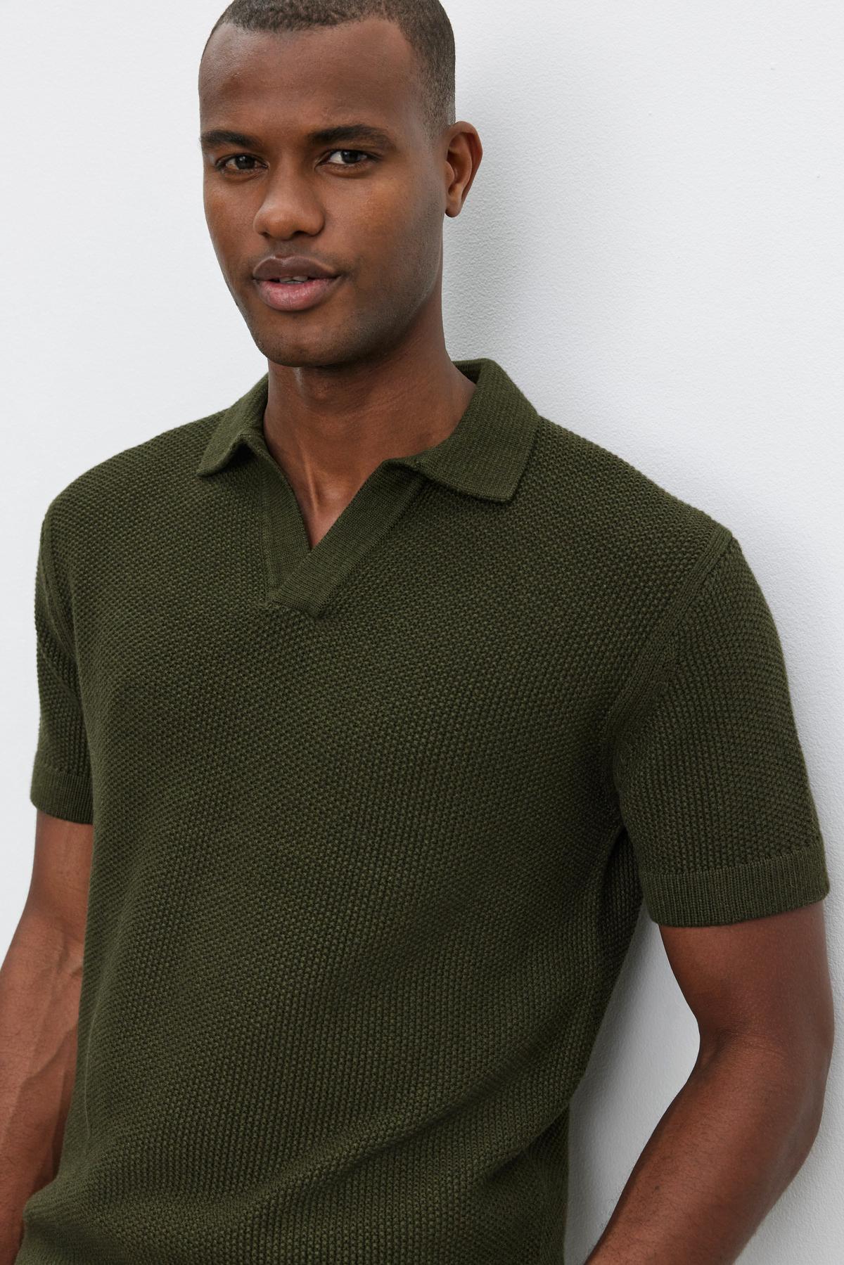   A man wearing the NIKLAS SWEATER POLO by Velvet by Graham & Spencer, a dark green short-sleeve sweater polo with a subtle seed stitch texture, stands against a light-colored background. 