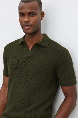 A man wearing the NIKLAS SWEATER POLO by Velvet by Graham & Spencer, a dark green short-sleeve sweater polo with a subtle seed stitch texture, stands against a light-colored background.