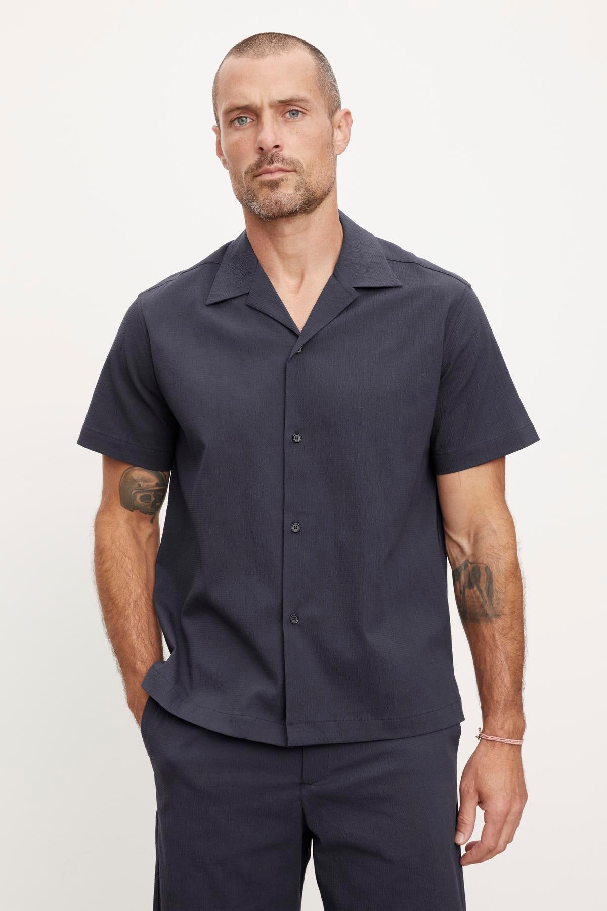   A man with a neutral expression wearing a dark grey relaxed fit short-sleeved Velvet by Graham & Spencer FRANK BUTTON-UP SHIRT and matching pants, showing visible tattoos on both arms. 
