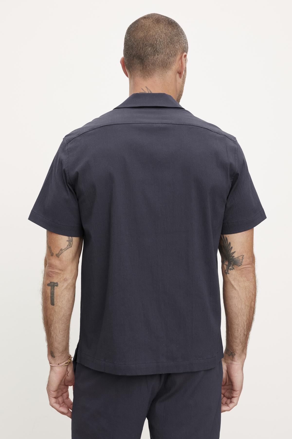   Rear view of a man wearing a dark grey short-sleeved seersucker cotton Velvet by Graham & Spencer shirt and pants, showing tattoos on his arms. 