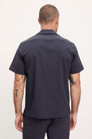Rear view of a man wearing a dark grey short-sleeved seersucker cotton Velvet by Graham & Spencer shirt and pants, showing tattoos on his arms.