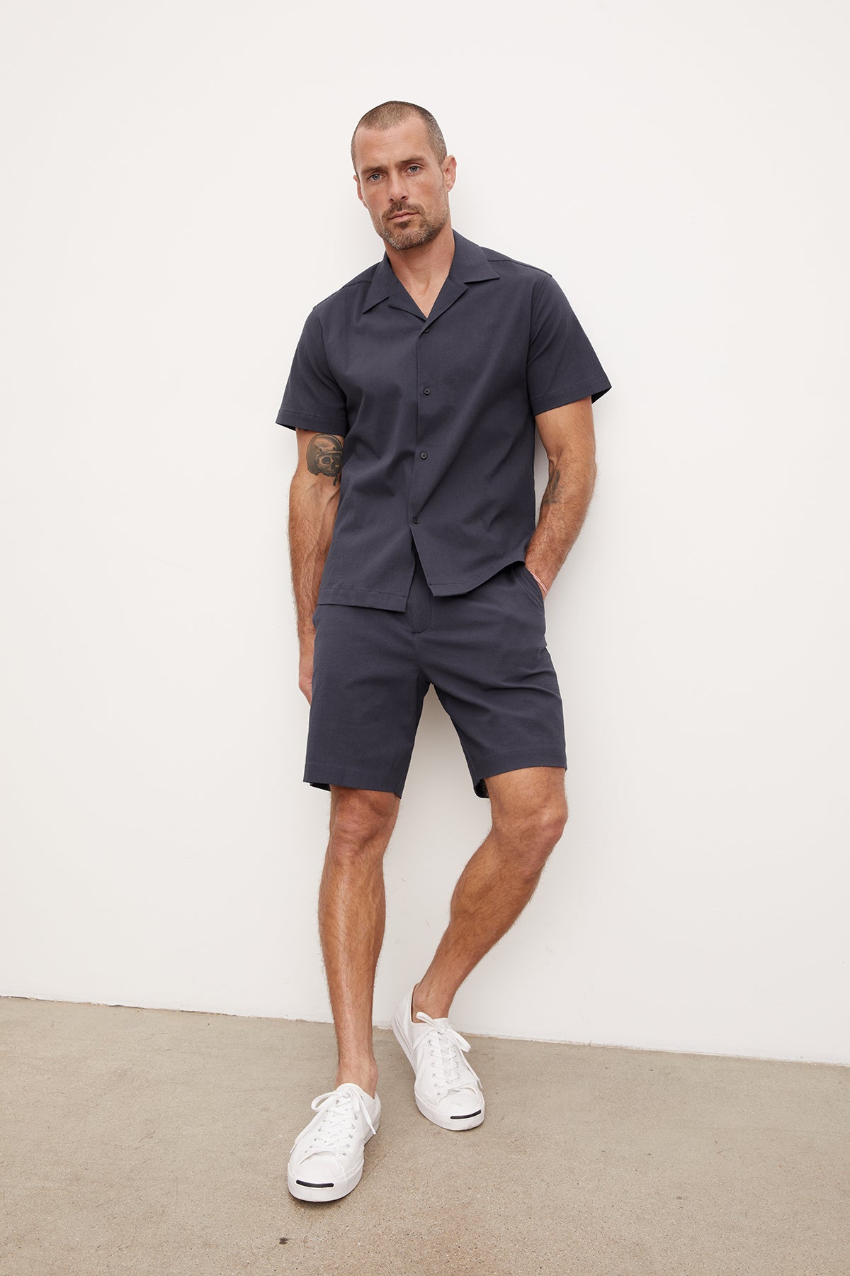   A man in a navy blue seersucker cotton FRANK BUTTON-UP SHIRT and matching shorts stands against a white background. He wears white sneakers and has a tattoo on his left arm. (Brand Name: Velvet by Graham & Spencer) 