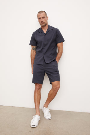 A man in a navy blue seersucker cotton FRANK BUTTON-UP SHIRT and matching shorts stands against a white background. He wears white sneakers and has a tattoo on his left arm. (Brand Name: Velvet by Graham & Spencer)