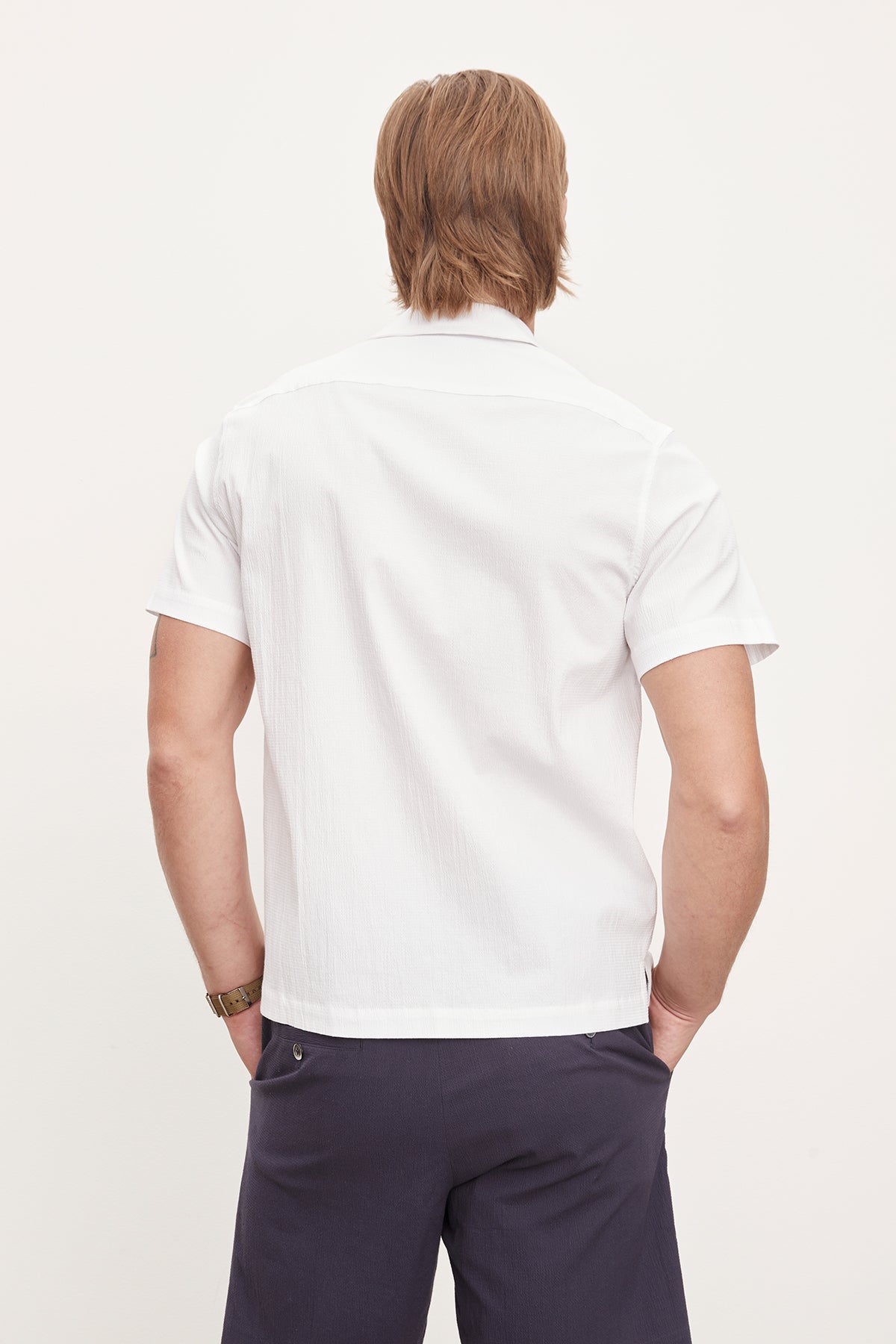   Man standing with his back to the camera, wearing a white seersucker cotton FRANK BUTTON-UP SHIRT by Velvet by Graham & Spencer and dark trousers, against a plain light background. 