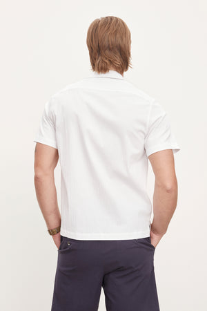 Man standing with his back to the camera, wearing a white seersucker cotton FRANK BUTTON-UP SHIRT by Velvet by Graham & Spencer and dark trousers, against a plain light background.