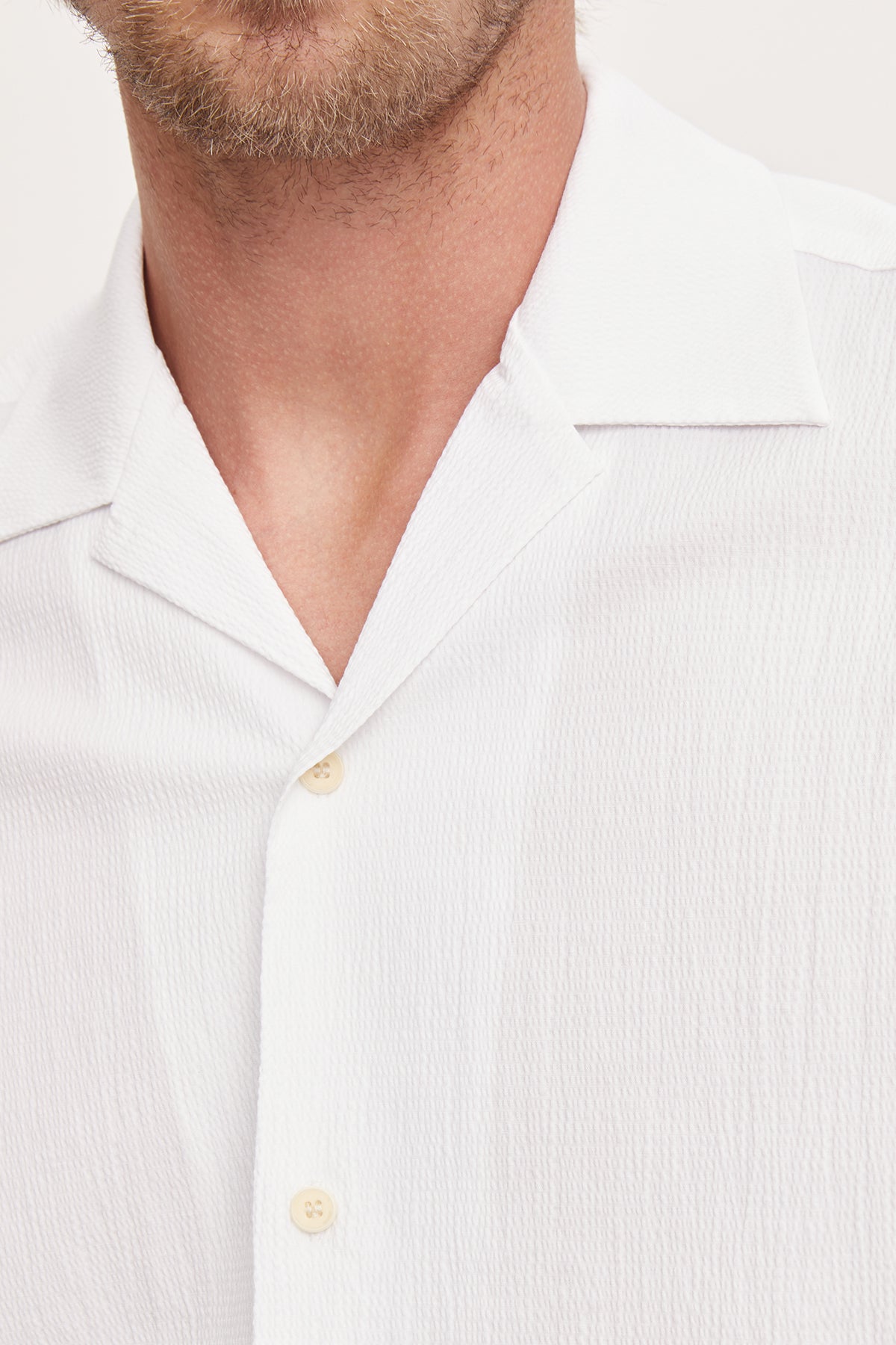   Close-up of a man wearing a white seersucker cotton Velvet by Graham & Spencer FRANK button-up shirt with the collar slightly open, showing two buttons. 