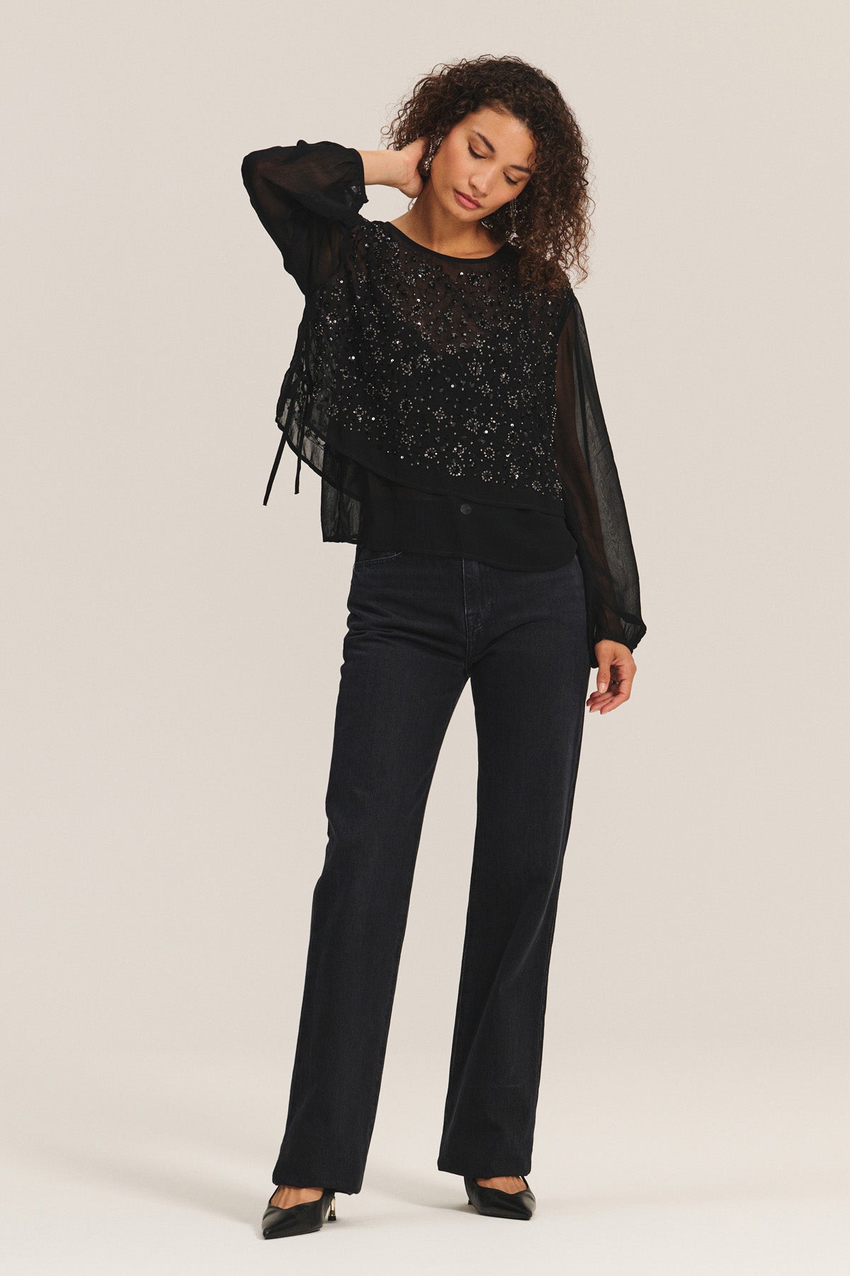 A woman stands against a plain background wearing the MARLOWE TOP by Velvet by Graham & Spencer— a sheer black cotton-viscose top with embellishments, paired with dark jeans and black heels for an effortlessly chic holiday look.