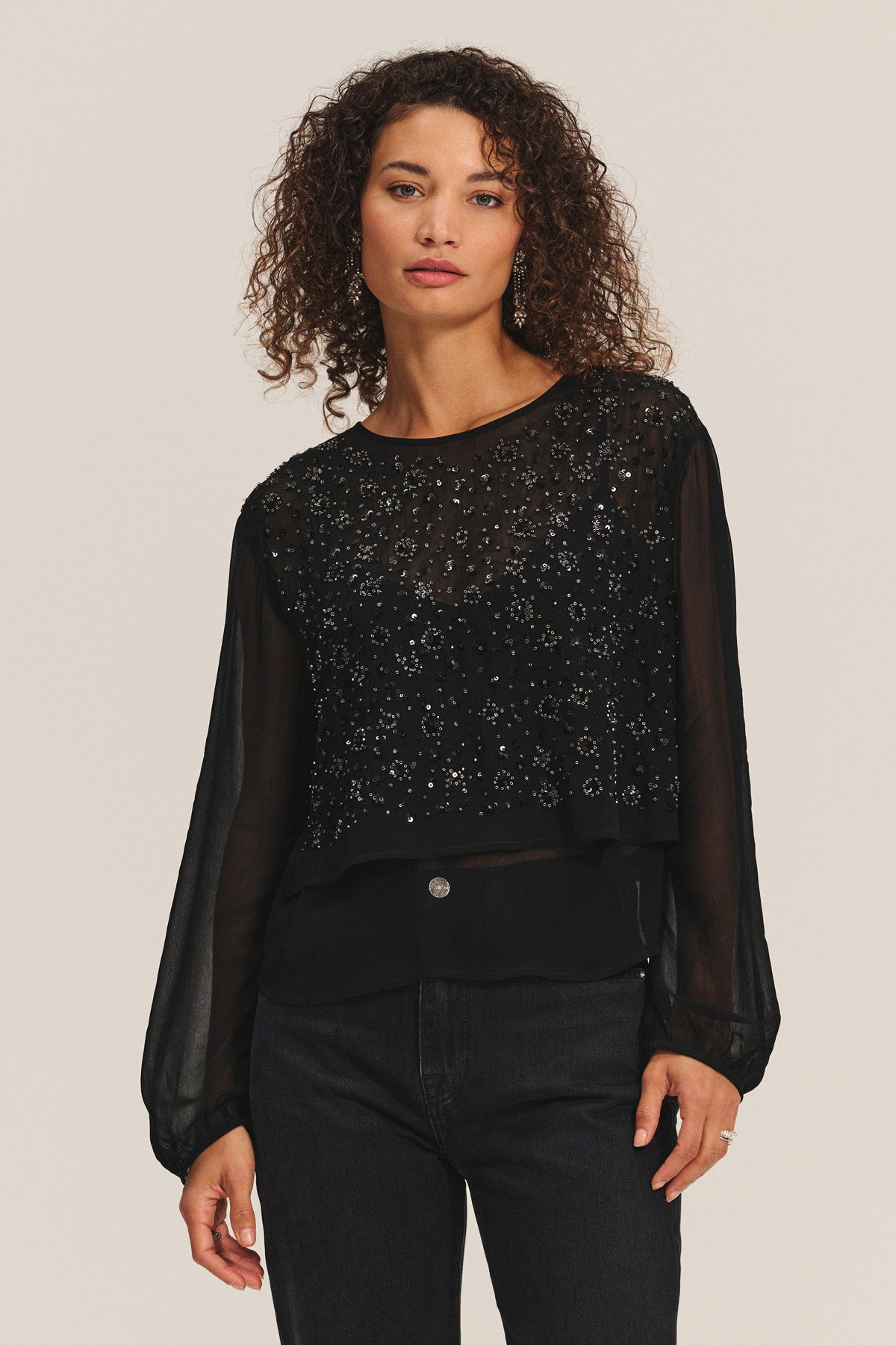 A woman with curly hair wears the MARLOWE TOP by Velvet by Graham & Spencer— a sheer cotton-viscose blouse with beaded embellishments—paired with black jeans, standing against a plain light background.-40263654604993