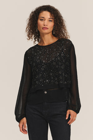 A woman with curly hair wears the MARLOWE TOP by Velvet by Graham & Spencer— a sheer cotton-viscose blouse with beaded embellishments—paired with black jeans, standing against a plain light background.