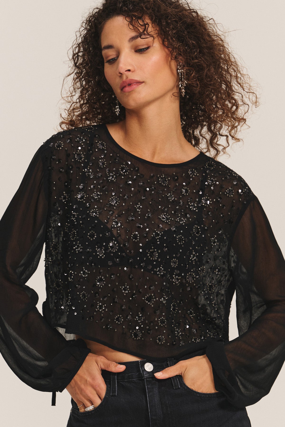 A woman with curly hair wears the MARLOWE TOP by Velvet by Graham & Spencer— a sheer cotton-viscose blouse adorned with sequins and beads, styled with high-waisted dark jeans, creating a stunning holiday party look against a plain backdrop.