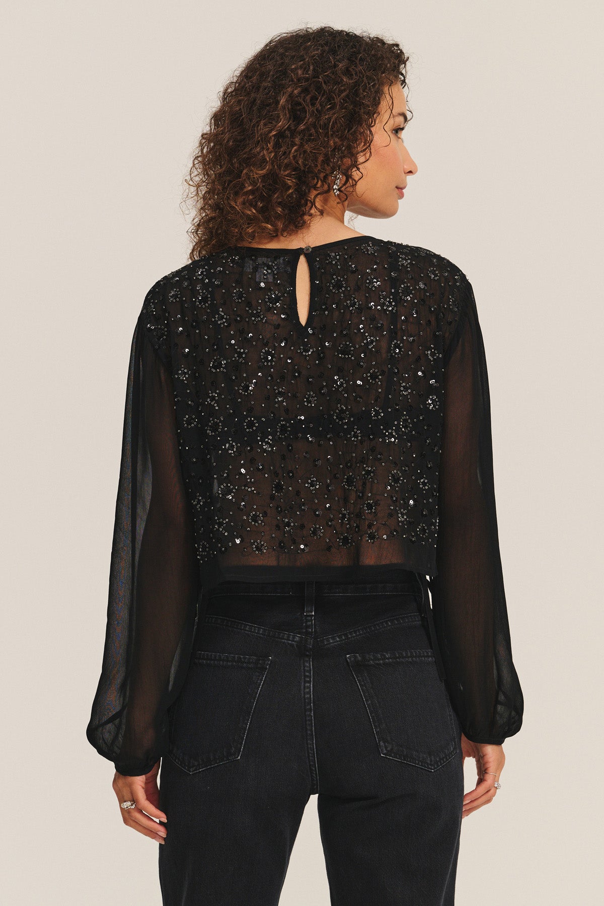 Woman with curly hair shown from the back in the MARLOWE TOP by Velvet by Graham & Spencer—a sheer cotton-viscose piece with black beading—paired with high-waisted black jeans, against a light background; perfect for holiday parties.