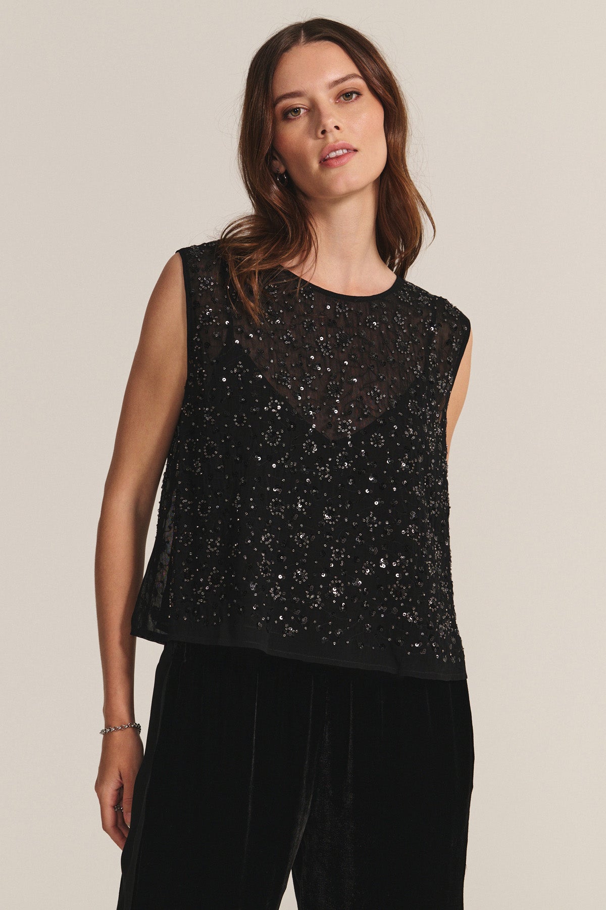A woman stands against a plain light background wearing the HILMA PANT from Velvet by Graham & Spencer, styled with a sleeveless black top featuring sequins and sheer details.