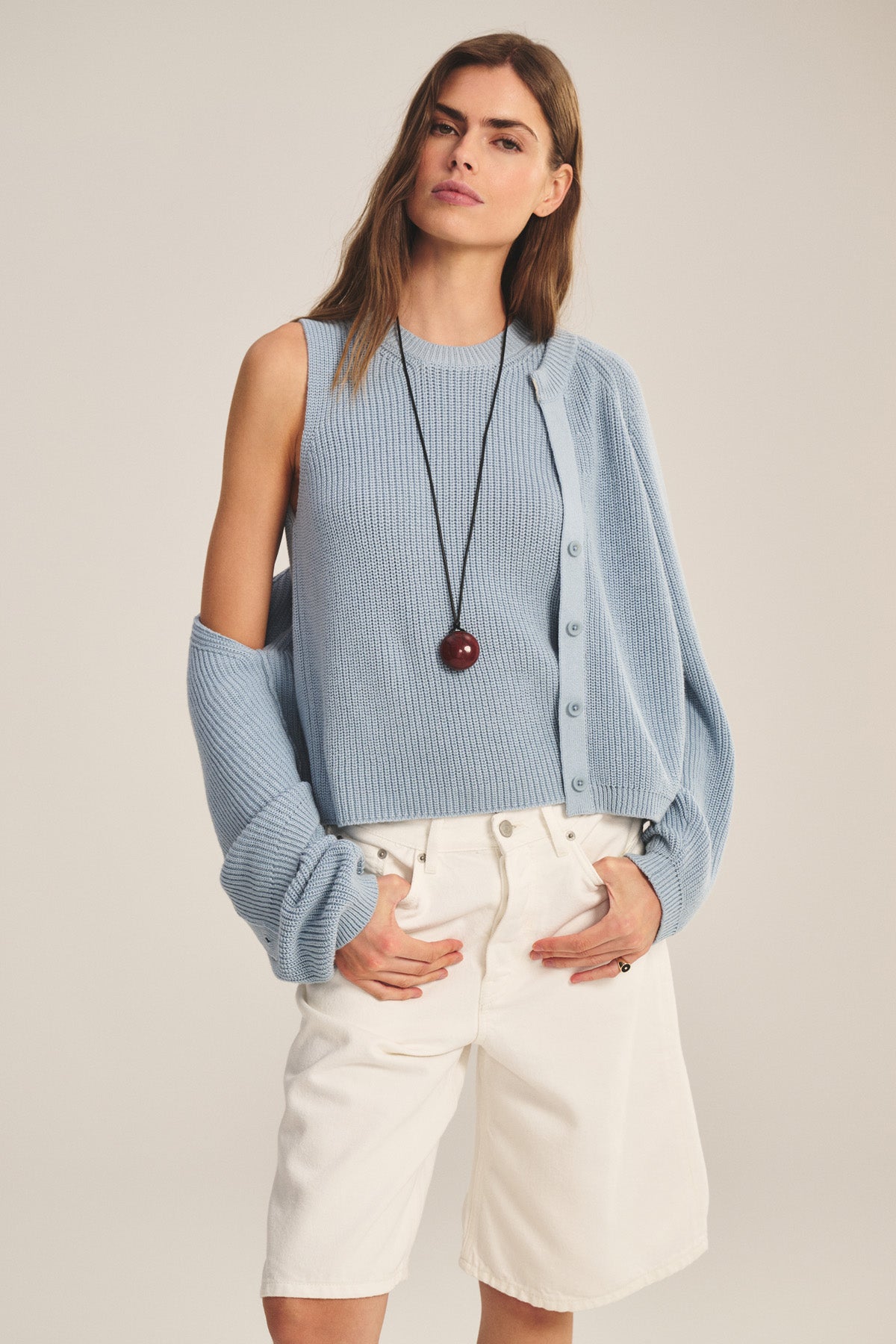 A woman wears the SERETTA SWEATER TANK by Velvet by Graham & Spencer, a sleeveless light blue Pima cotton piece, paired with white knee-length shorts, a long round pendant necklace, and carries a matching cardigan.-40909904117953