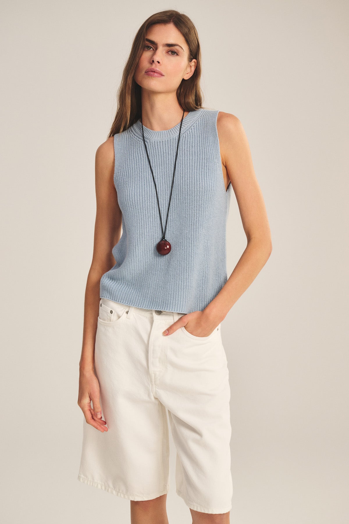 A woman wears the SERETTA SWEATER TANK by Velvet by Graham & Spencer in light blue, paired with white knee-length shorts and a long necklace with a round pendant, standing against a plain background.