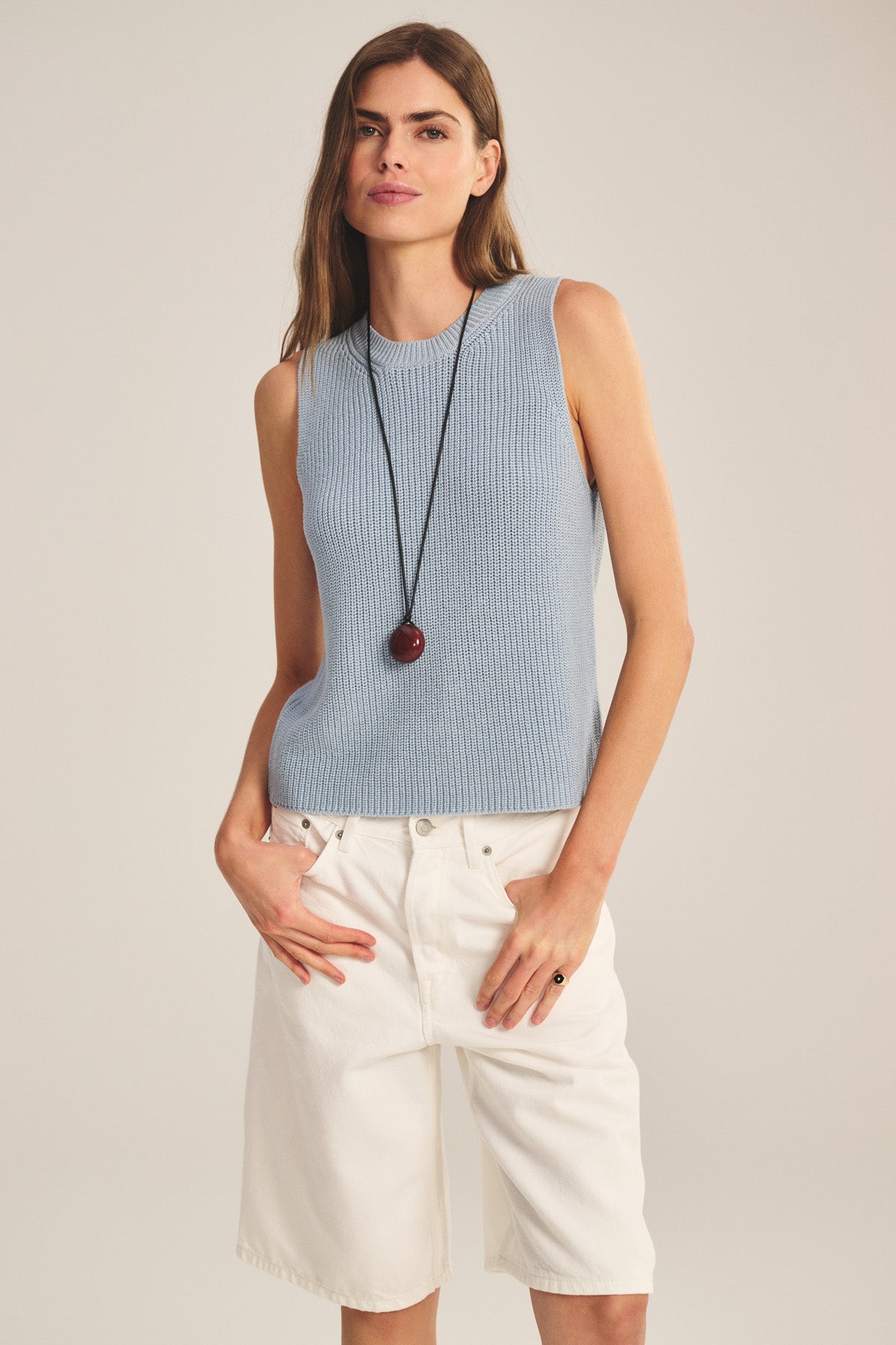 A woman wears the Velvet by Graham & Spencer SERETTA SWEATER TANK, a sleeveless light blue top with a ribbed crew neckline, paired with white knee-length shorts and a long round pendant necklace.-40910121435329