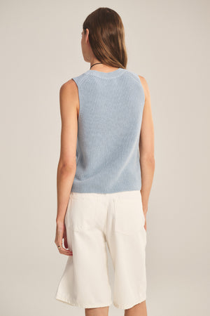 A person with long brown hair faces away, wearing the Velvet by Graham & Spencer SERETTA SWEATER TANK in light blue with a ribbed crew neckline and white knee-length shorts against a neutral background.