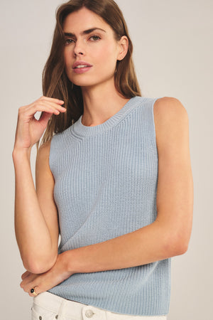 A woman wears the SERETTA SWEATER TANK by Velvet by Graham & Spencer—a light blue, ribbed crew neck, sleeveless top—paired with white pants, standing against a plain background.