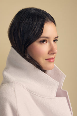 A woman with dark hair wears the ABBY BLAZER by Velvet by Graham & Spencer—a light-colored wool blazer featuring a high collar and oversized silhouette—gazing to the right against a plain beige background, showcasing an effortlessly layered look for everyday style.