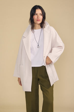 A woman models the ABBY BLAZER by Velvet by Graham & Spencer—an oversized, blazer-style white coat—layered over a white t-shirt with green pants and a necklace, standing against a plain beige background for a chic take on everyday layering.