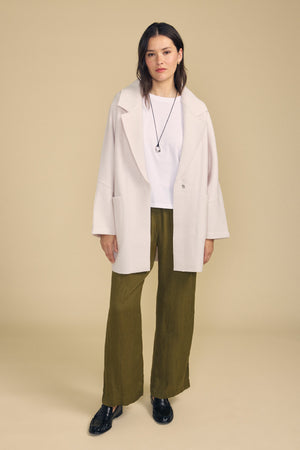 A woman stands against a beige background wearing the ABBY BLAZER by Velvet by Graham & Spencer—an oversized white wool blazer styled with a white shirt, olive green wide-leg pants, black shoes, and a black necklace—perfect for everyday layering.