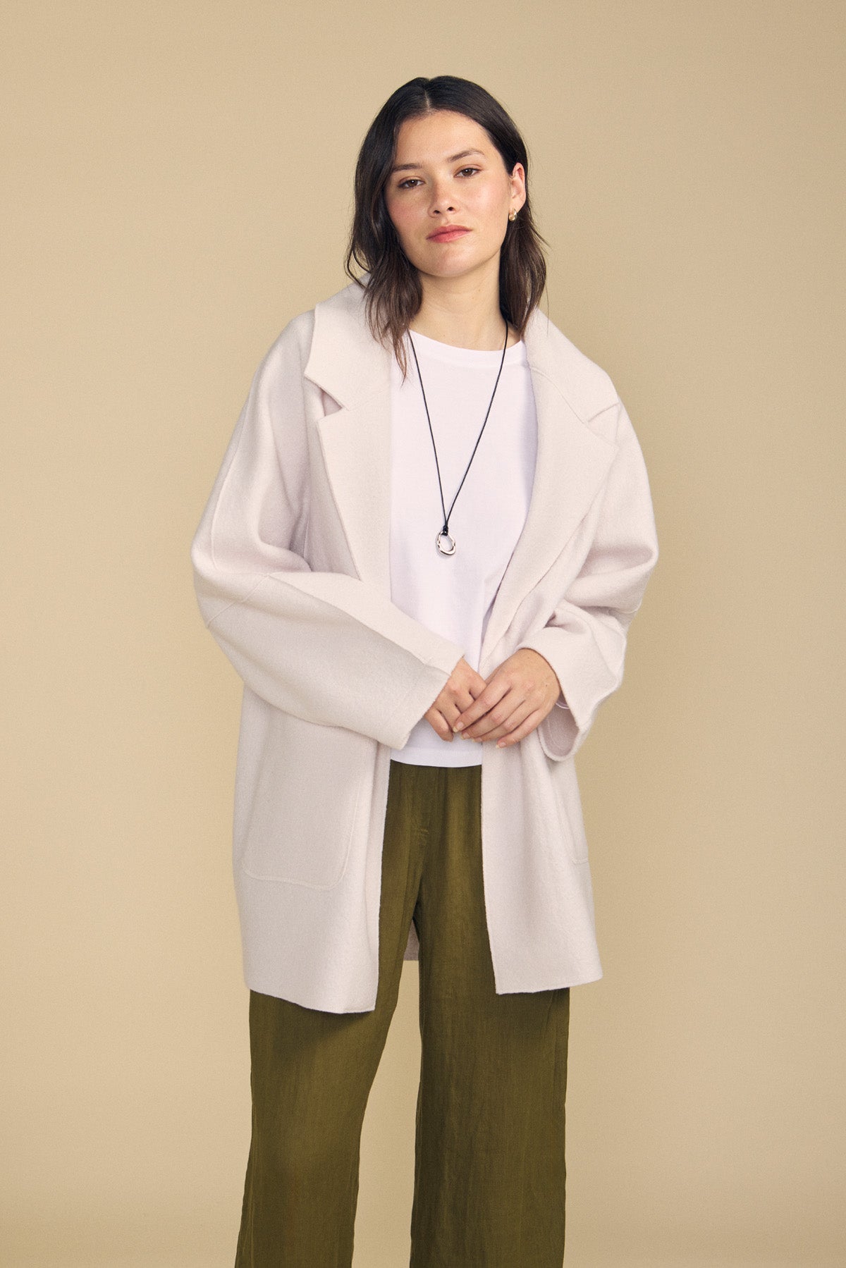 A woman stands against a beige background wearing the ABBY BLAZER by Velvet by Graham & Spencer—a white wool blazer featuring an oversized silhouette—layered over a white shirt, paired with olive green pants and a black necklace for effortlessly chic everyday style.-41860415488193