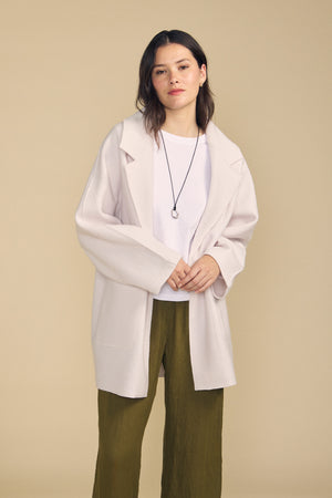 A woman stands against a beige background wearing the ABBY BLAZER by Velvet by Graham & Spencer—a white wool blazer featuring an oversized silhouette—layered over a white shirt, paired with olive green pants and a black necklace for effortlessly chic everyday style.