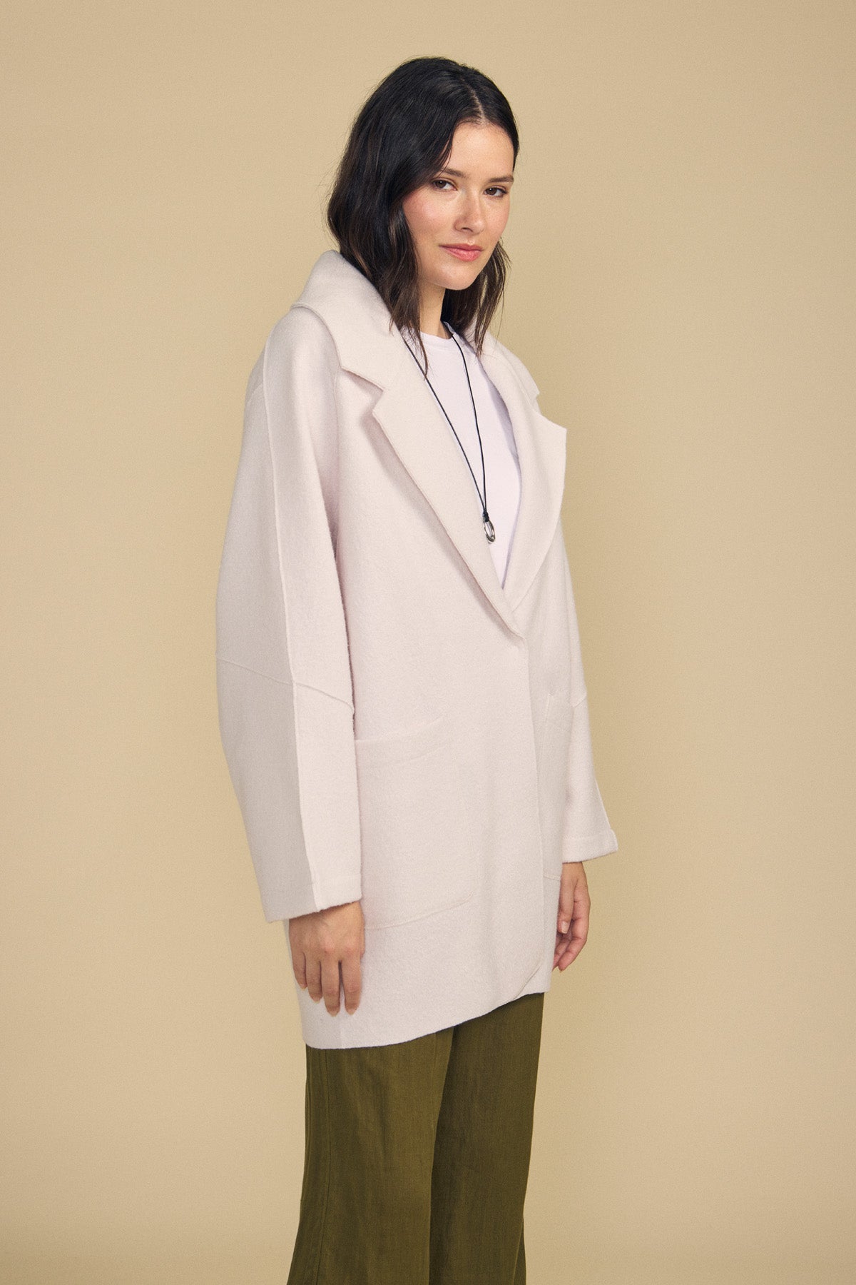 A woman wears the ABBY BLAZER by Velvet by Graham & Spencer—an oversized, light beige coat—layered over a white top and green pants, accessorized with a necklace, and stands against a plain beige background. Perfect for everyday layering.