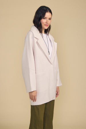 A woman wears the ABBY BLAZER by Velvet by Graham & Spencer—an oversized, light beige coat—layered over a white top and green pants, accessorized with a necklace, and stands against a plain beige background. Perfect for everyday layering.