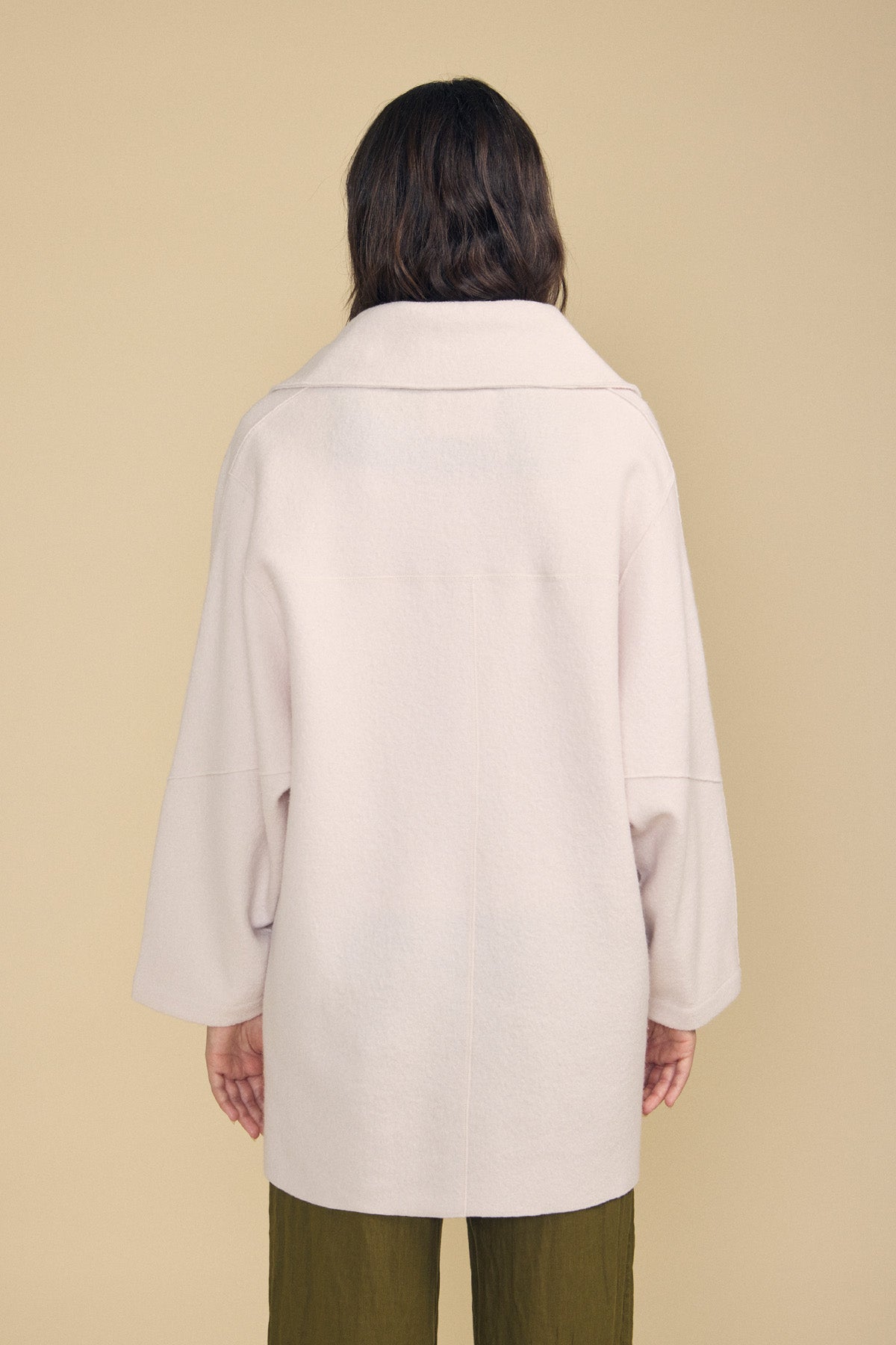 A person with long brown hair stands facing away from the camera against a beige background, embodying an effortless approach to everyday layering in Velvet by Graham & Spencer’s ABBY BLAZER, styled with olive green pants.