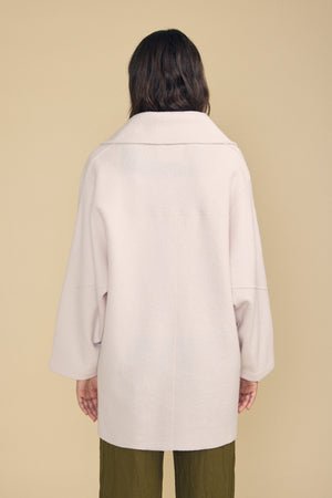 A person with long brown hair stands facing away from the camera against a beige background, embodying an effortless approach to everyday layering in Velvet by Graham & Spencer’s ABBY BLAZER, styled with olive green pants.