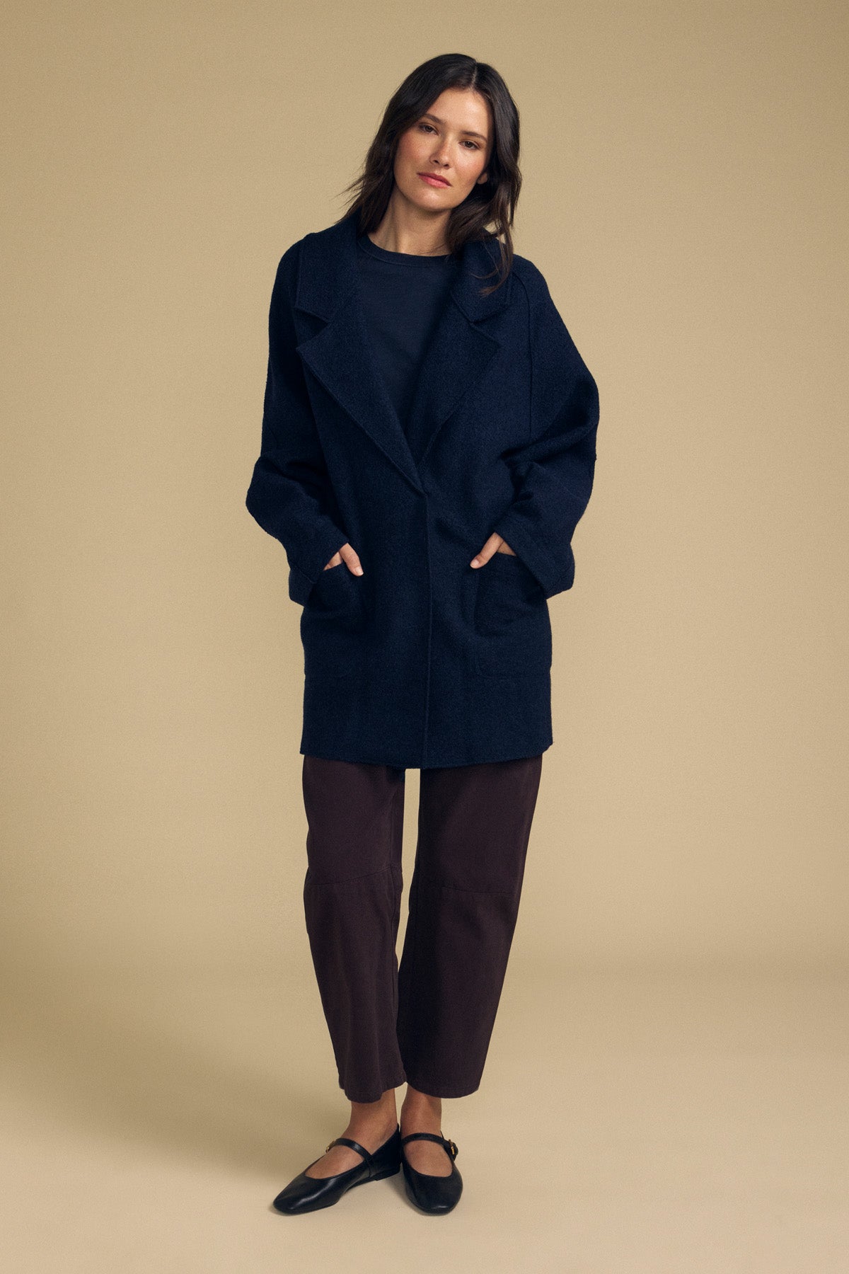 A woman stands against a beige background wearing the ABBY BLAZER by Velvet by Graham & Spencer—an oversized dark wool blazer—styled with a black top, cropped brown pants, and black flats, her hands in the coat pockets for an effortlessly layered everyday look.