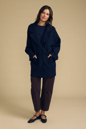 A woman stands against a beige background wearing the ABBY BLAZER by Velvet by Graham & Spencer—an oversized dark wool blazer—styled with a black top, cropped brown pants, and black flats, her hands in the coat pockets for an effortlessly layered everyday look.